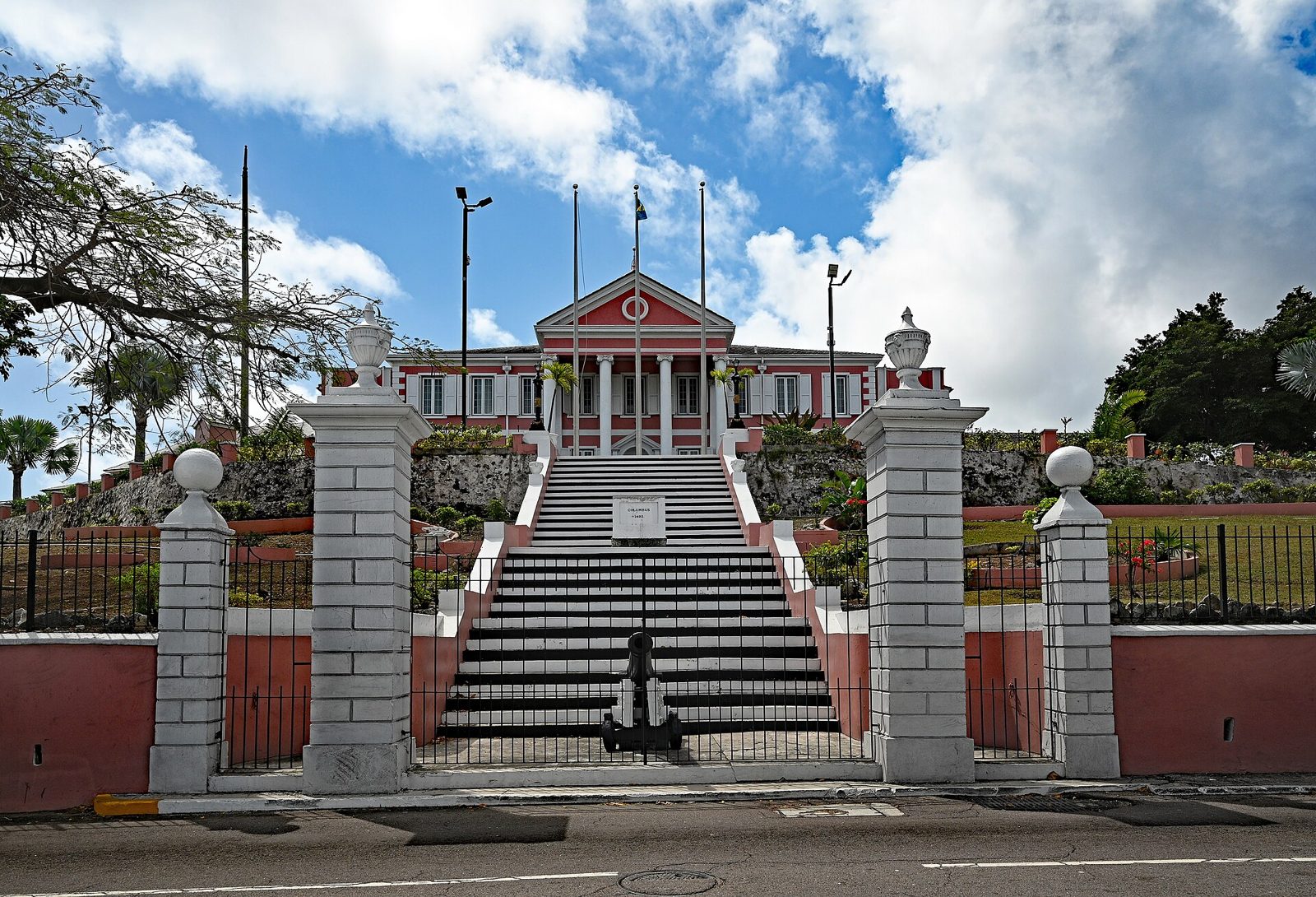 Pink colonial mansion with white columns at the top of a long flight of stone steps behind a gate, under a blue sky with clouds