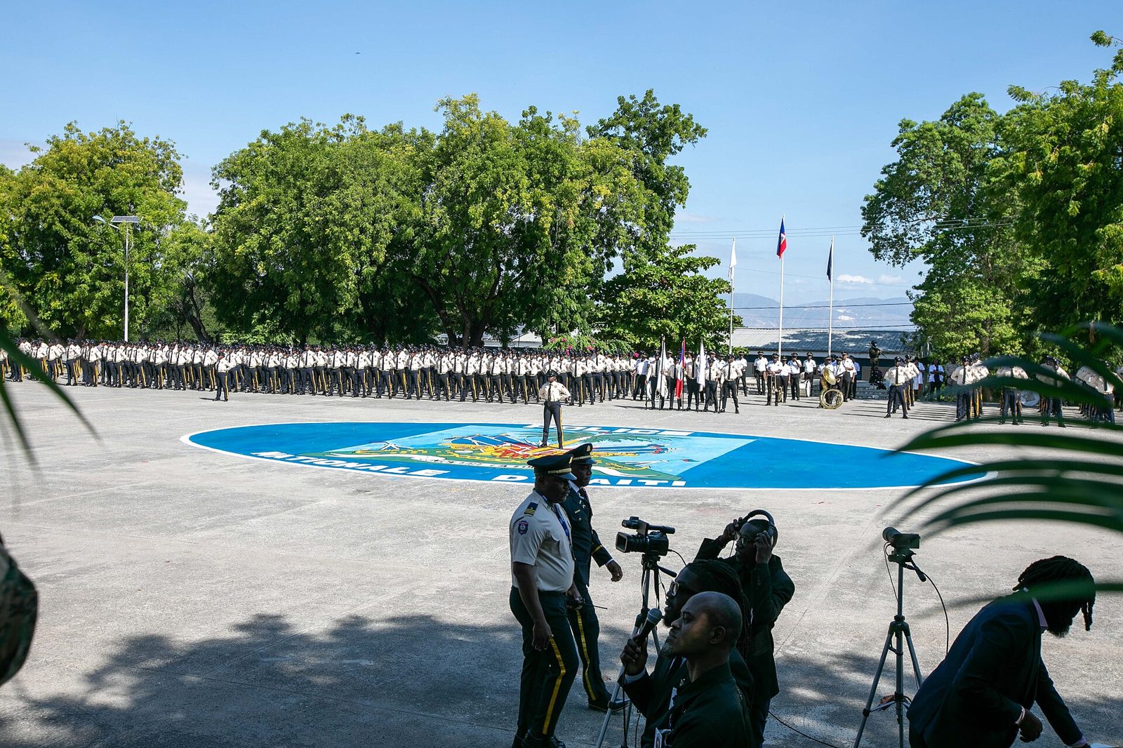 Rows of uniformed police recruits standing in formation on a large paved parade ground with trees behind and camera crews in the foreground