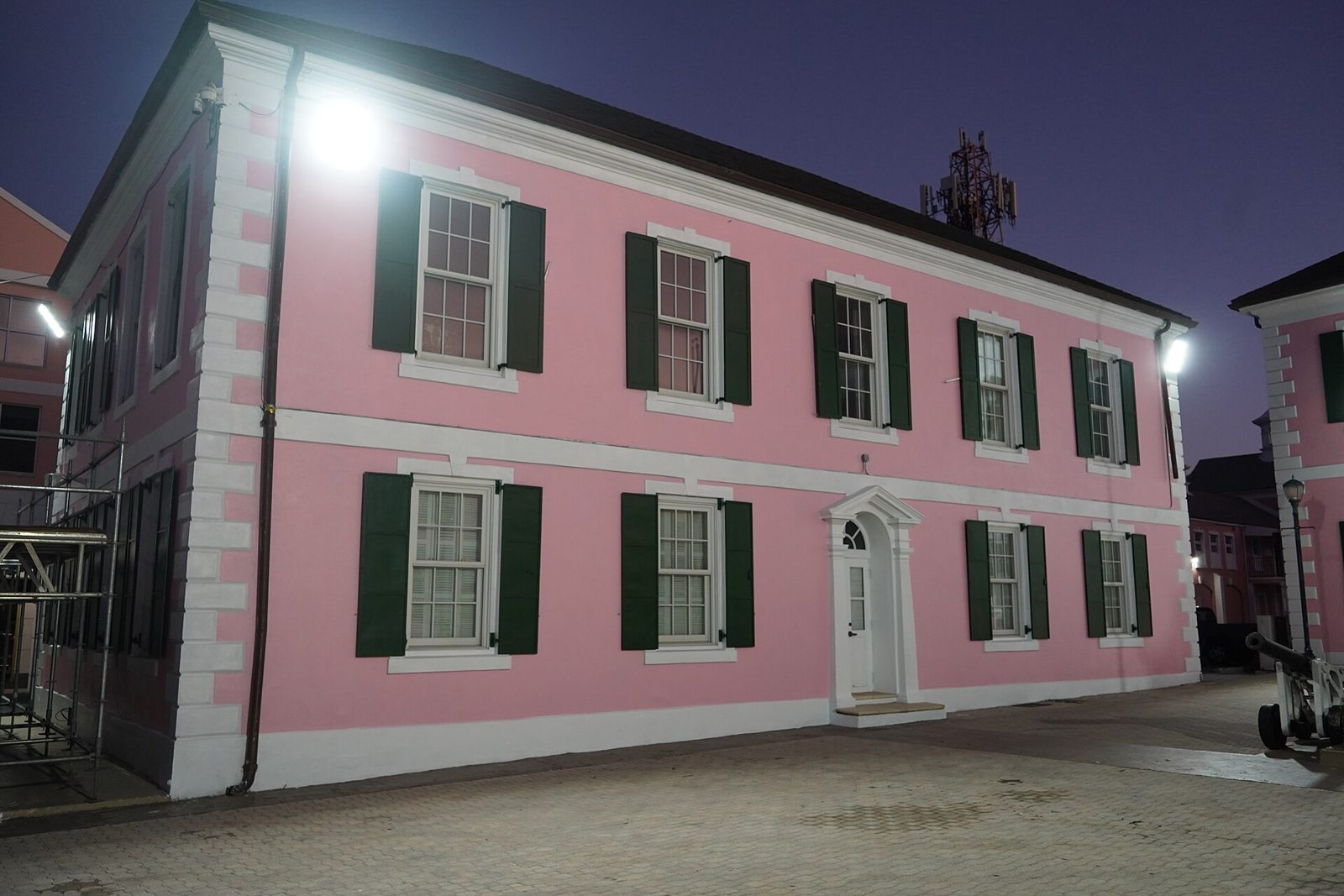 Two-storey pink colonial building with dark green shutters and white trim, lit up at dusk on a paved square in Nassau