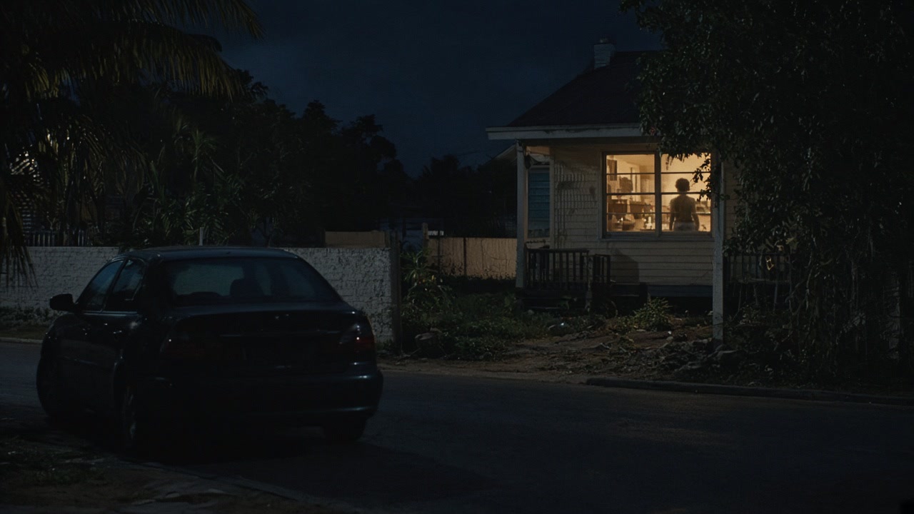 A parked car across from a lit window where a woman sits alone