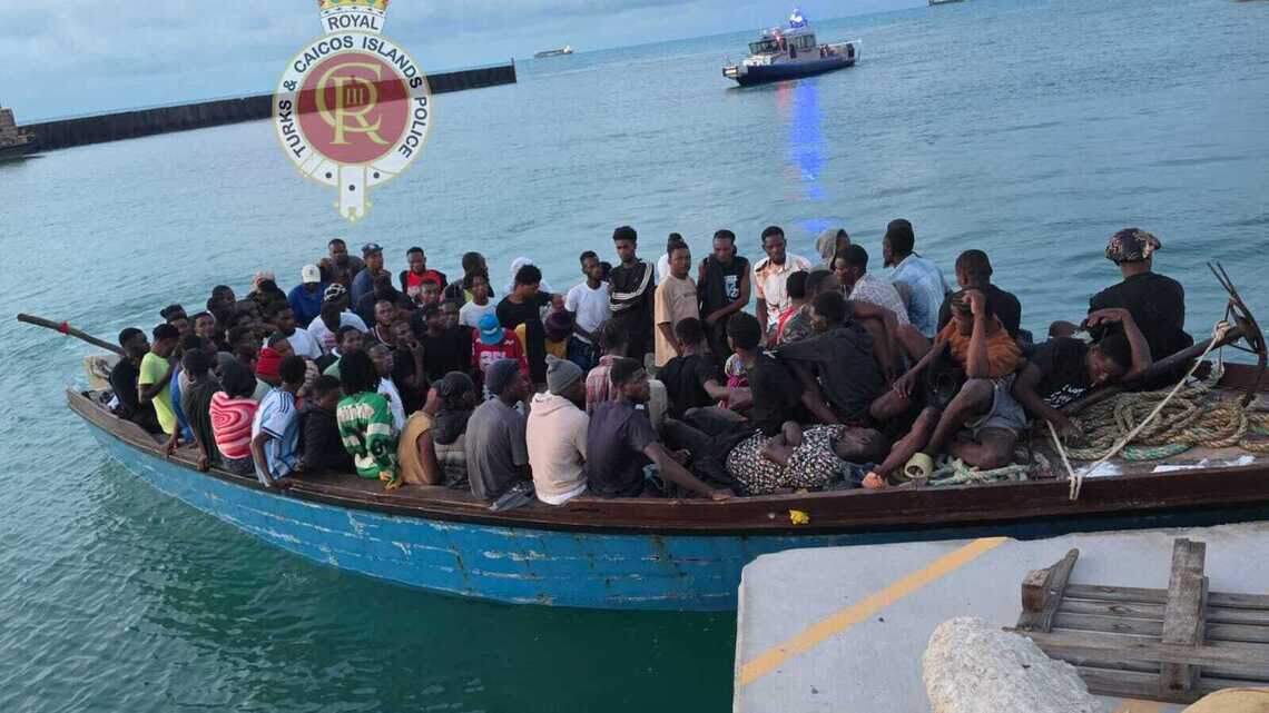 Wooden migrant sloop packed with passengers beside a dock, with a police vessel behind it